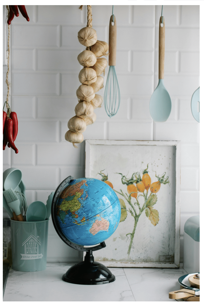 Kitchen counter with globe, botanical art, herbs and spices, and pastel utensils against a white tile backsplash.