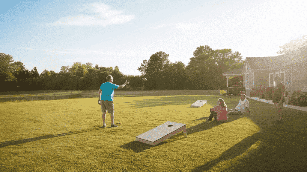 a backyard lawn game area with a cornhole set placed on a flat grassy yard with people enjoying the game on a sunny afternoon