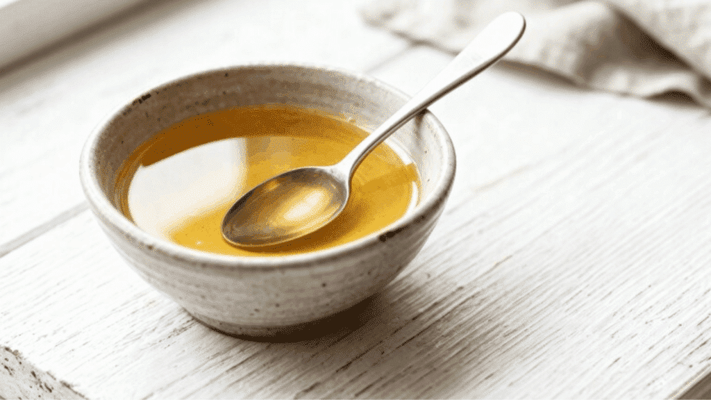 a ceramic bowl filled with warm melted coconut oil and a silver spoon resting inside placed on a white wooden surface with a linen cloth in the background