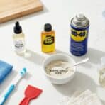 a flat lay of cleaning essentials baby oil, Goo Gone, WD-40, dish soap, baking soda paste, a microfiber cloth, a toothbrush, and a red scraper on a white counter