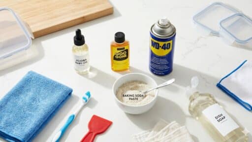 a flat lay of cleaning essentials baby oil, Goo Gone, WD-40, dish soap, baking soda paste, a microfiber cloth, a toothbrush, and a red scraper on a white counter