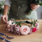 a floral crown made of pink and purple flowers sits on a wooden table, with a person assembling a bouquet in the background.