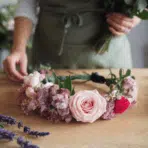 a floral crown made of pink and purple flowers sits on a wooden table, with a person assembling a bouquet in the background.