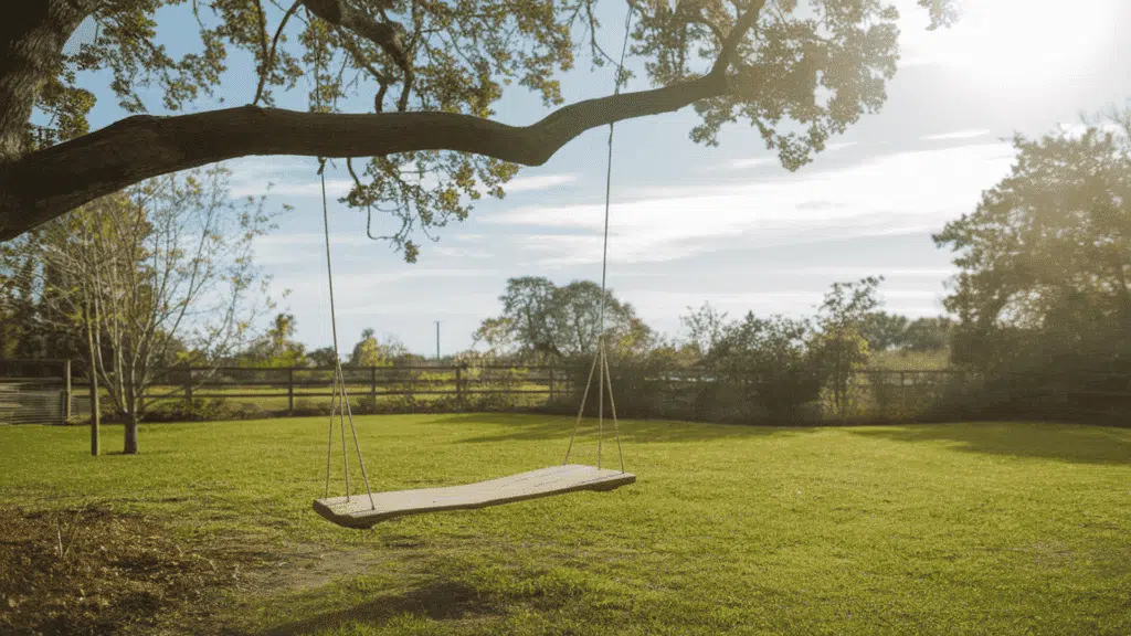 a simple wooden tree swing hanging from a thick strong branch in a lush green backyard on a bright and pleasant sunny afternoon