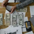 Person counting cash at wooden desk with financial documents, laptop, and smartphone calculator