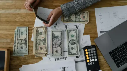 Person counting cash at wooden desk with financial documents, laptop, and smartphone calculator