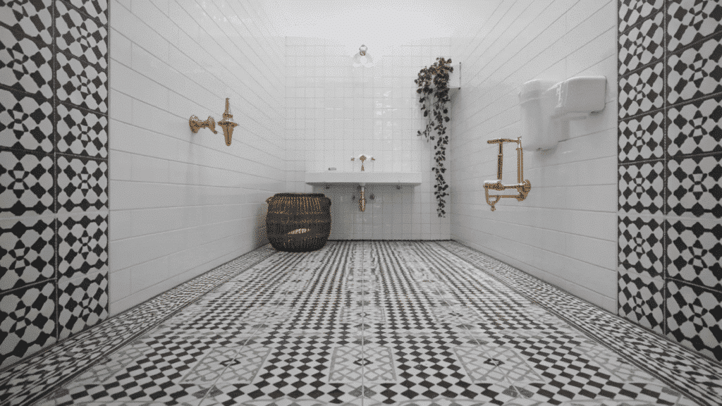 black and white bathroom with patterned encaustic floor tiles, white walls, brass fixtures, rattan basket, and plant highlighting the floor.