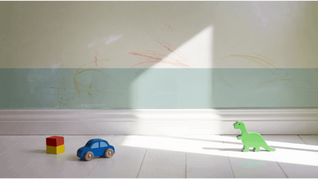 children's room wall with crayon marks and scribbles on eggshell painted surface with small toys on the floor in natural sunlight