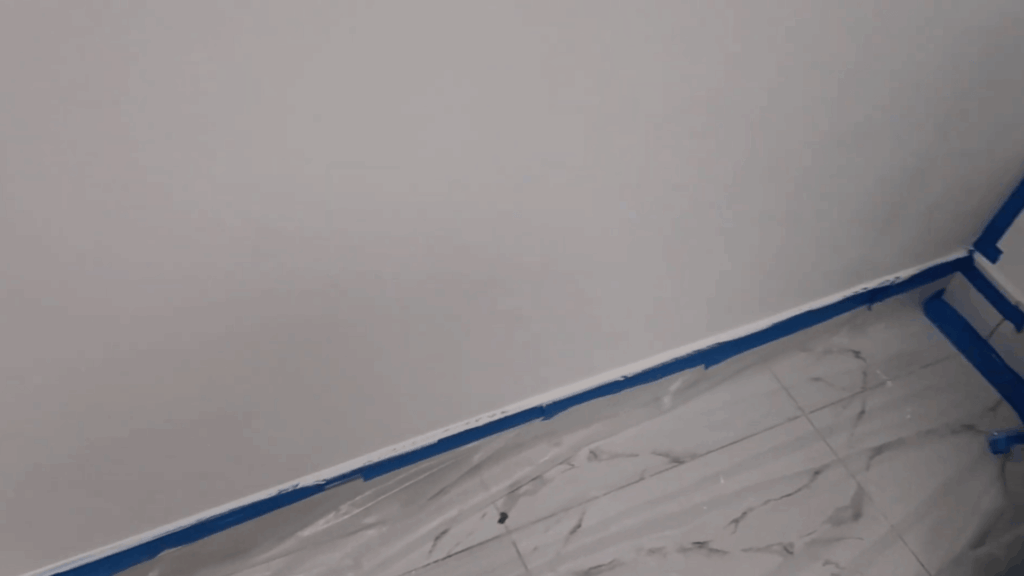 corner of a room with white wall and baseboard taped with blue painter's tape, plastic covering marble floor for painting prep