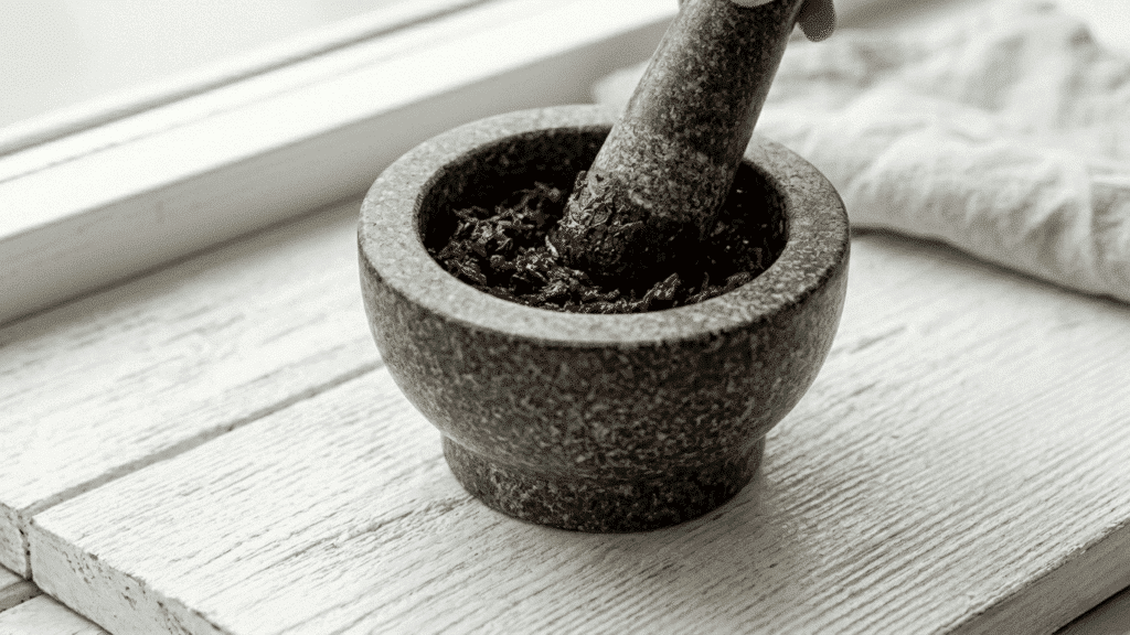 fresh hibiscus petals being ground into a dark paste in a stone mortar and pestle placed on a white wooden surface with a linen cloth beside it
