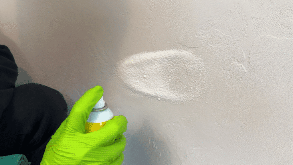 hand wearing green glove sprays cleaning foam onto a textured wall surface creating a white patch during wall cleaning process