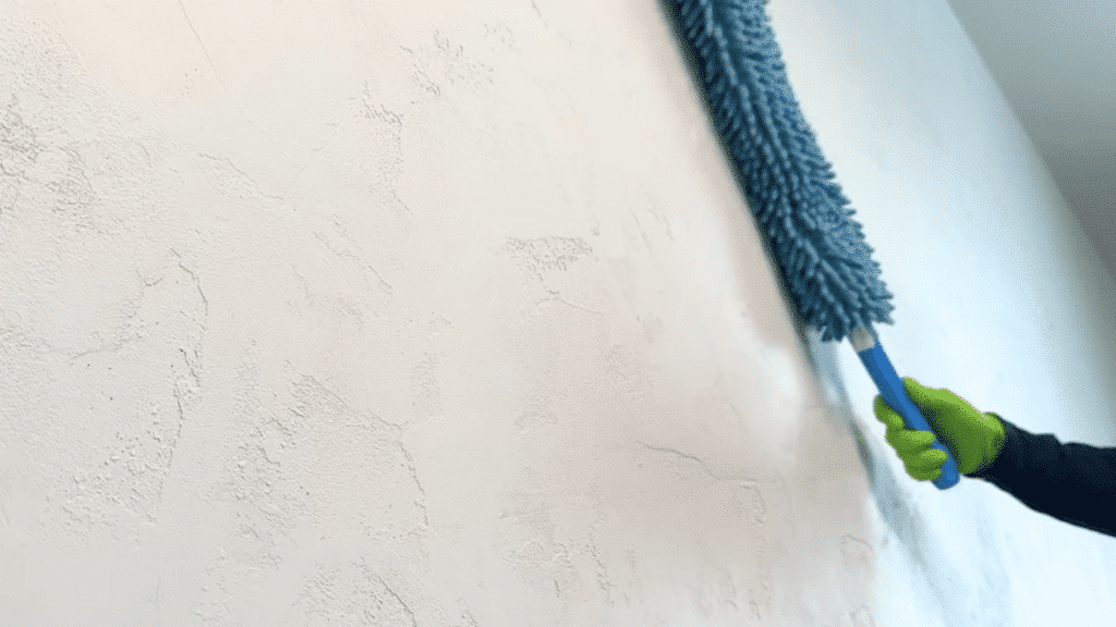 hand wearing green glove uses a blue handle duster to clean a textured white wall surface inside a room bright interior