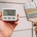 hands holding a digital thermometer and paintbrush near an exterior house wall checking the right temperature to paint outside