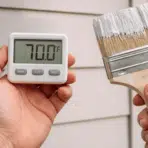 hands holding a digital thermometer and paintbrush near an exterior house wall checking the right temperature to paint outside