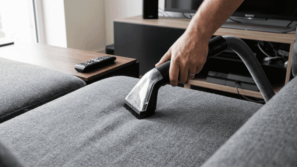 hands vacuuming sofa cushions and upholstery in a tidy modern living room during a routine cleaning session