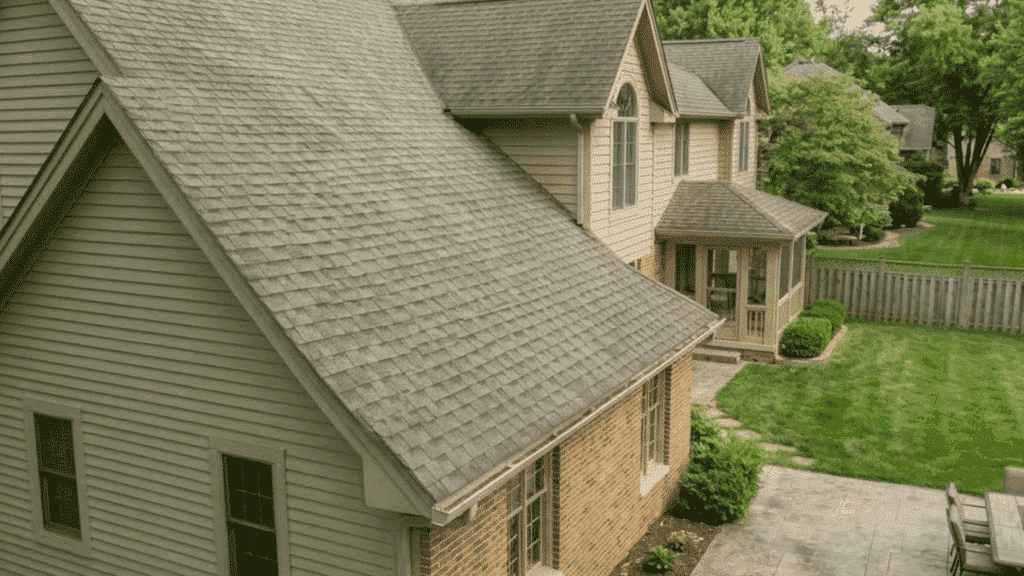 homeowner on ladder gently brushing dry brown moss off asphalt roof shingles after moss killer treatment under bright daylight (1)