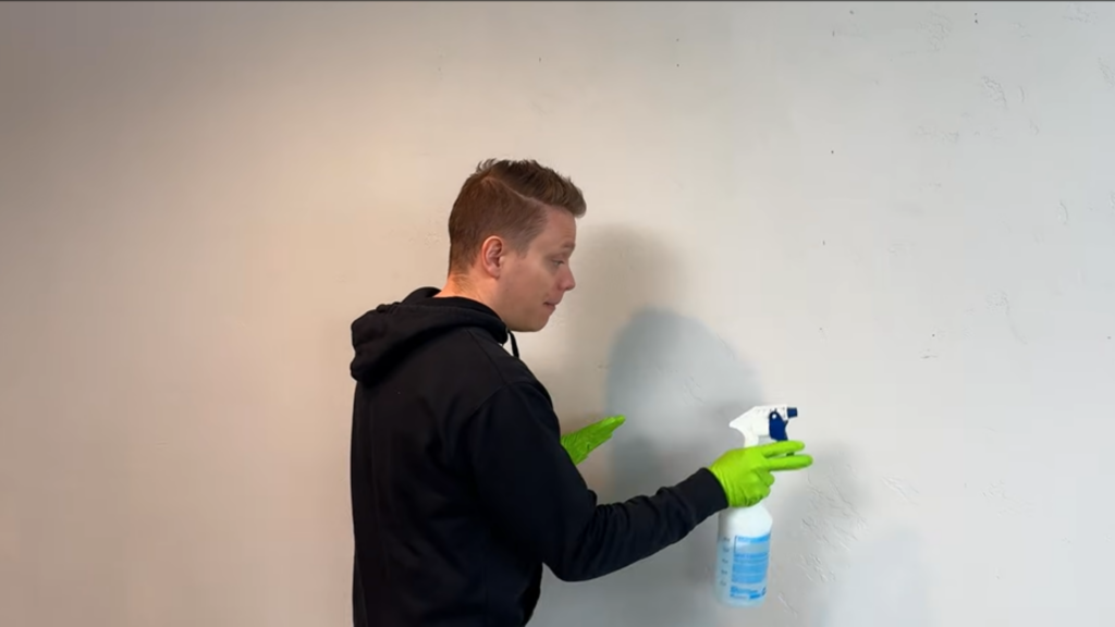 man wearing green gloves sprays cleaning solution onto a textured white wall using a spray bottle while standing indoors (1)