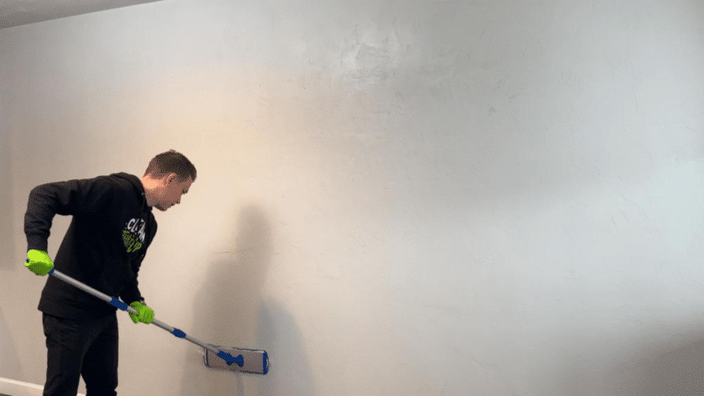 man wearing green gloves uses a flat mop to clean a textured white wall inside a room with cabinets and wood flooring visible (1)