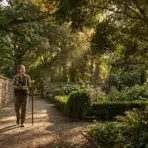 Surveyor with equipment walking through landscaped garden path under dappled sunlight