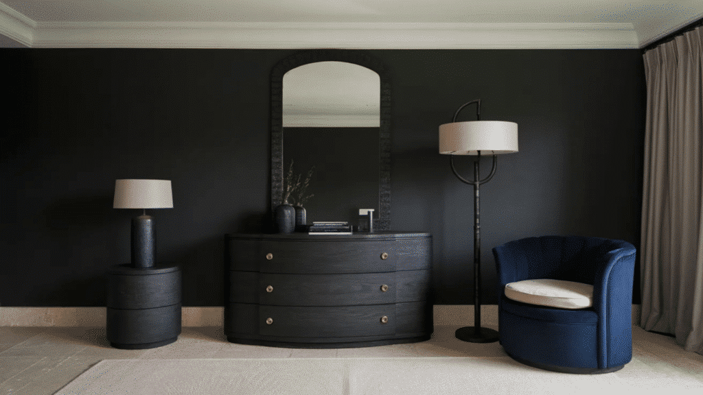 modern bedroom corner with black walls, curved dresser, arched mirror, floor lamp, and blue accent chair
