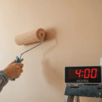painter applying a coat of interior wall paint with a roller beside a digital timer displaying 4 hours and a sign reading ready for second coat