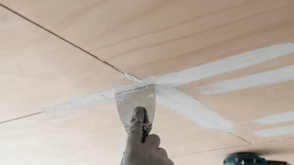 person applying joint compound to plywood ceiling seam with putty knife while a sander is nearby