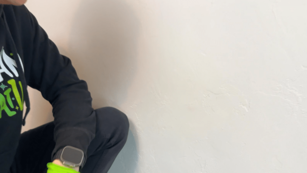 person in dark clothing and green gloves kneels near a clean textured white wall indoors after wiping surface during cleaning process