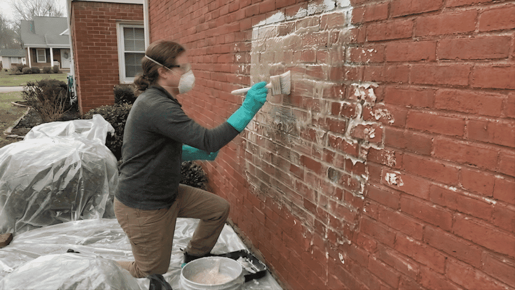 person in mask and teal gloves brushes a clear, glossy chemical gel onto a red brick wall