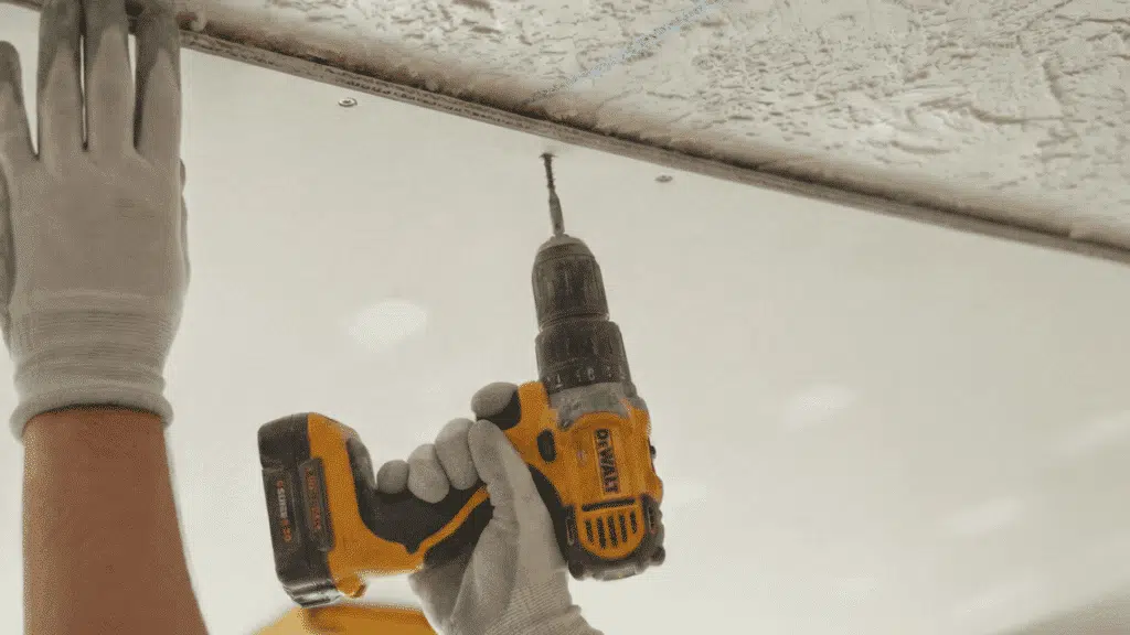 person installing drywall panel on ceiling using power drill and screws while wearing gloves