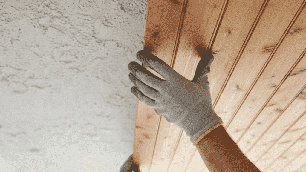 person installing natural wood tongue-and-groove planks on ceiling over textured surface while wearing gloves