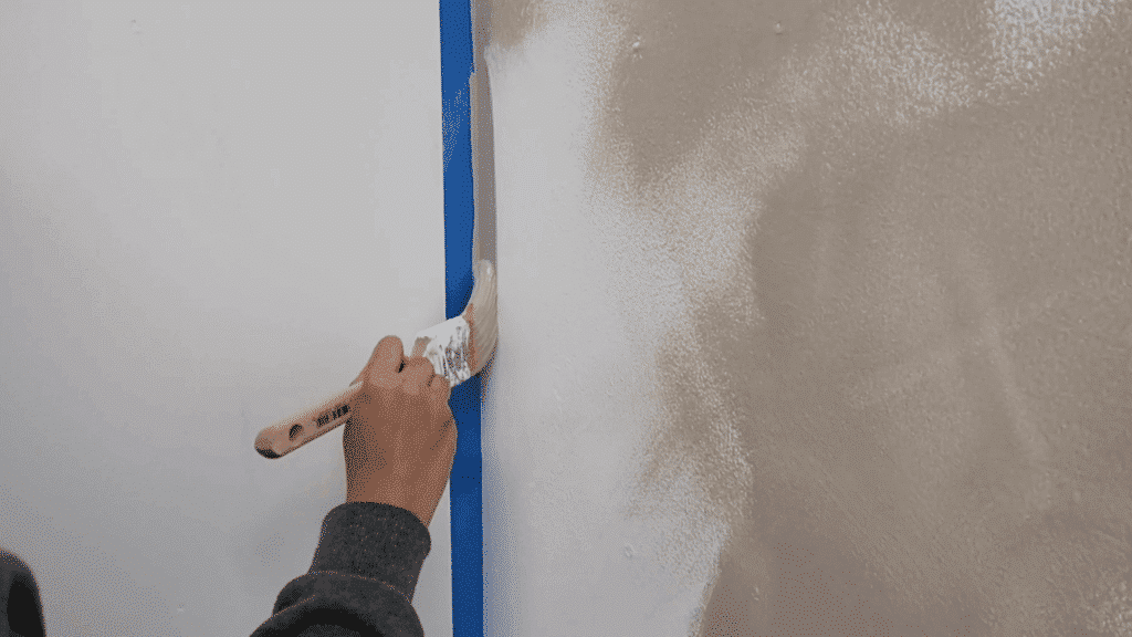 person painting wall edge with brush along blue painter’s tape, applying light beige paint next to white surface for clean line