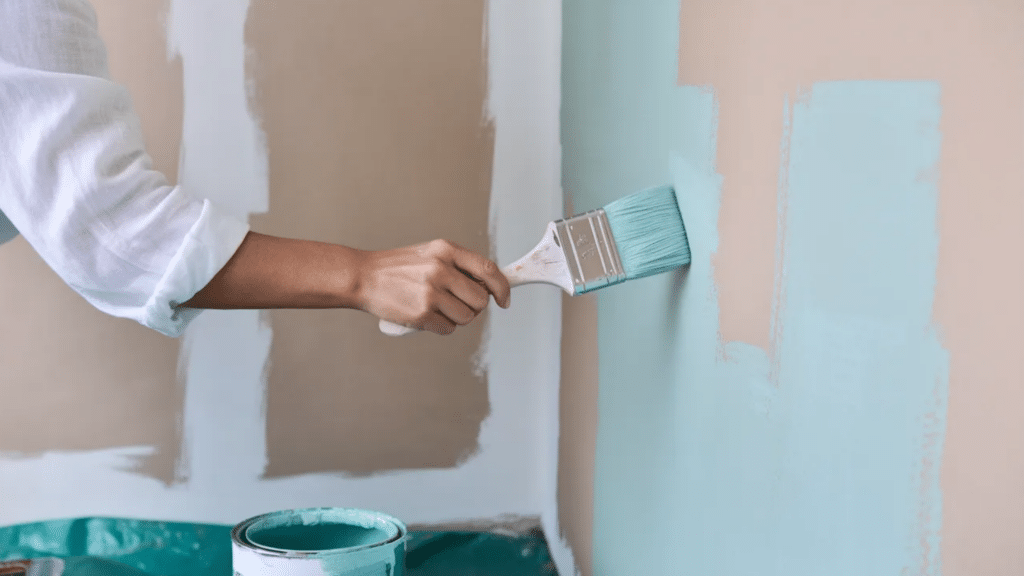 person using a paintbrush to apply light blue paint to an interior wall, with a paint can below and partially painted beige and white sections visible in the background