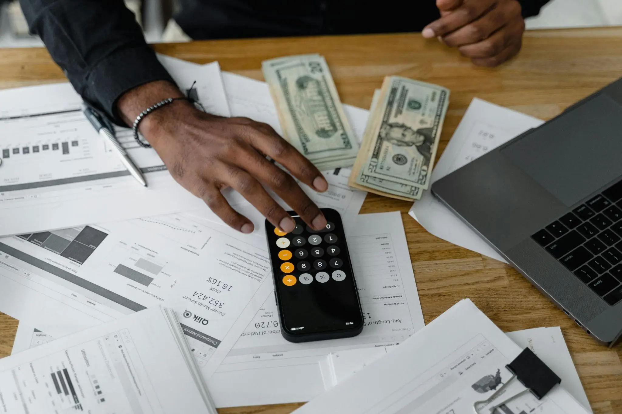 Hand using calculator on wooden desk with financial documents and stacks of cash