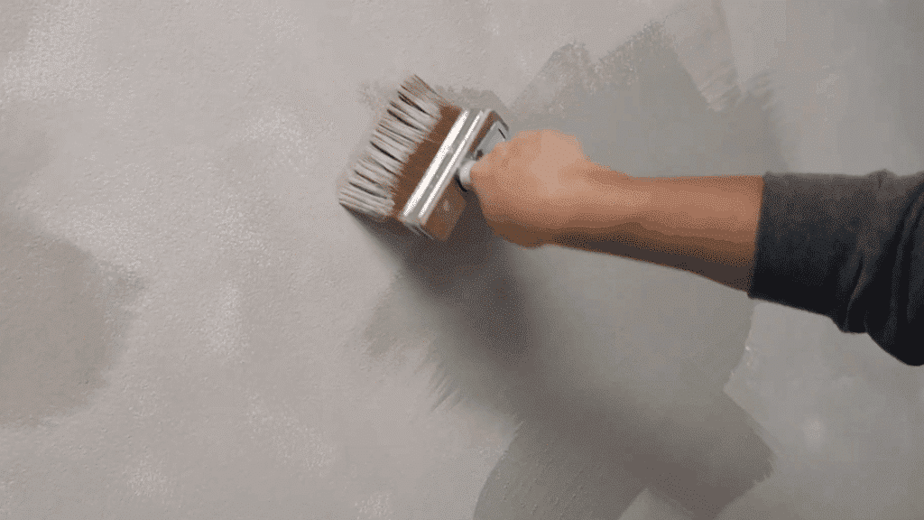 person using wide brush to apply textured beige paint on wall, creating layered finish with visible strokes and soft mottled effect