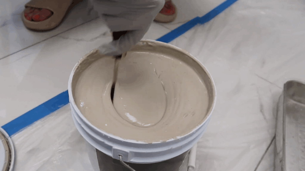 person wearing gloves stirring beige paint in a bucket on plastic-covered floor with blue tape, preparing surface for painting project