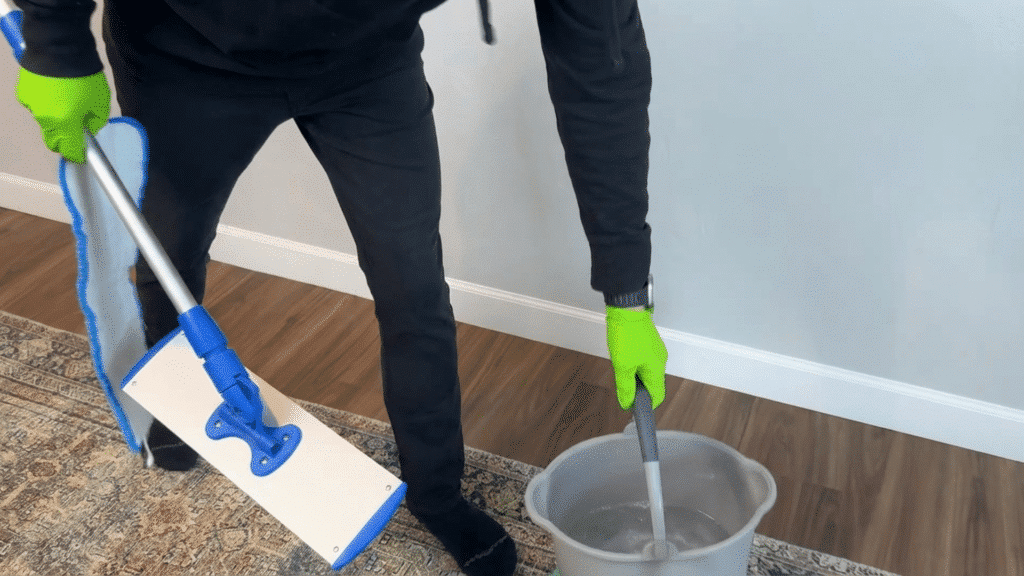 person wearing green gloves dips mop into bucket of water while holding flat mop head over rug on wood floor during cleaning