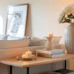 sofa table styled behind a grey sofa with a ceramic vase, book stack, candle tray, and framed art in a bright living room