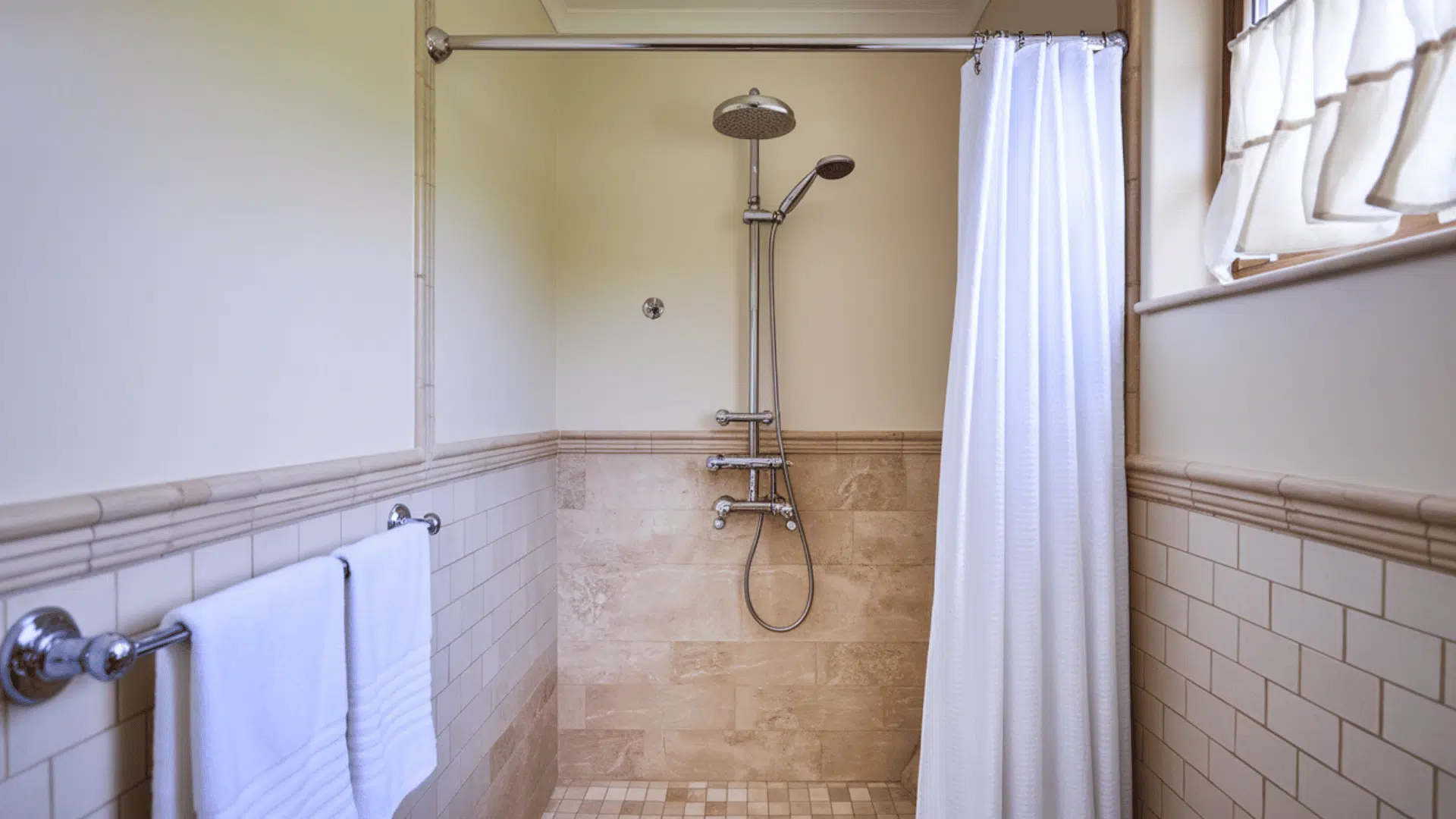 standard shower with white curtains, a chrome showerhead, and beige tiled walls in a simple bathroom setting.