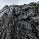 steep slate rock cliff with visible layers and fractures, showing natural weathering and moisture streaks on rugged surface