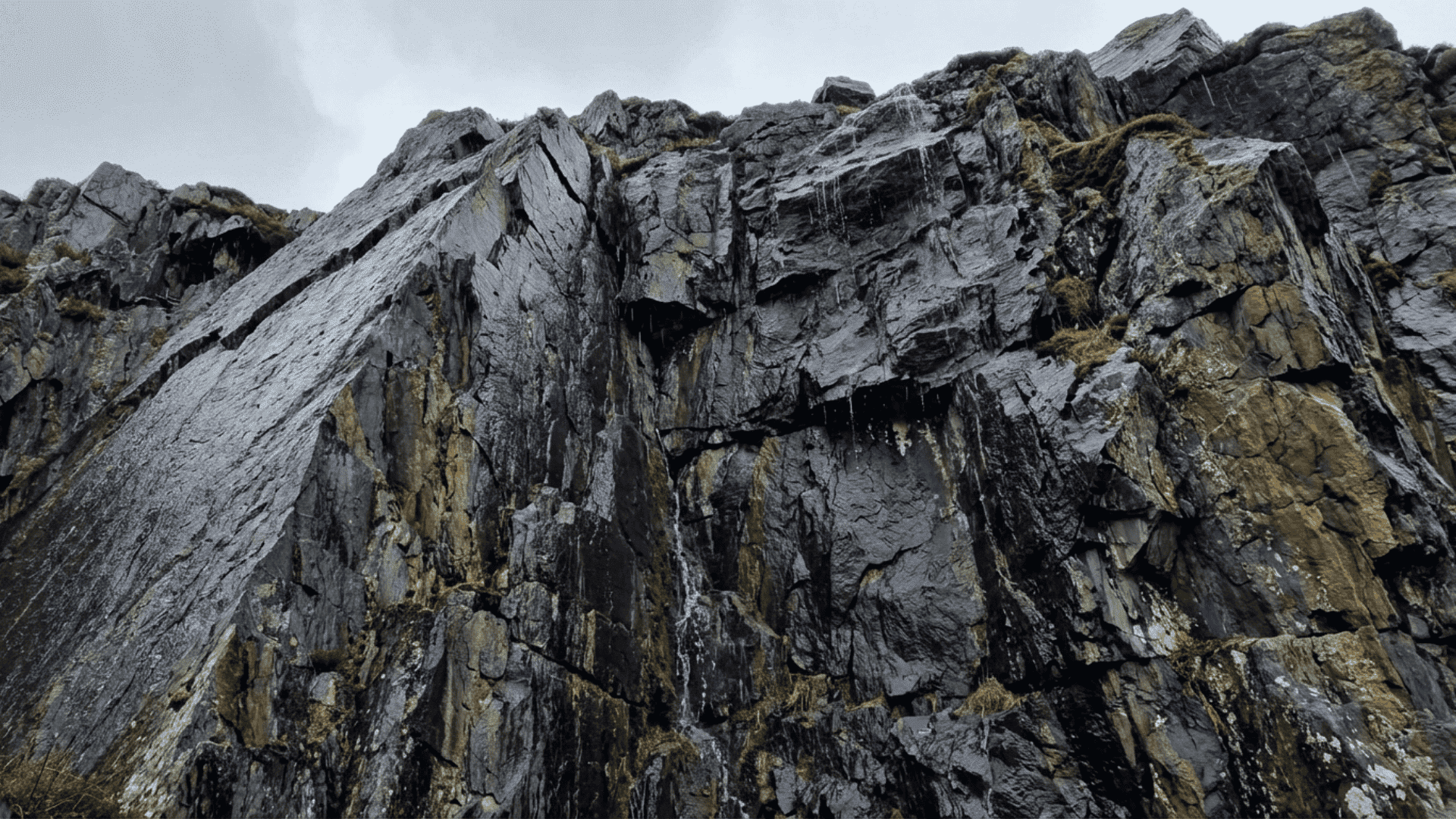 steep slate rock cliff with visible layers and fractures, showing natural weathering and moisture streaks on rugged surface