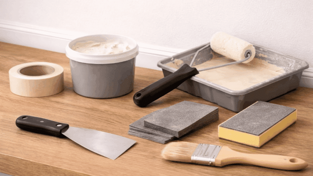 tools and materials like putty knife sandpaper, filler, primer, paint roller tray and cloth neatly arranged on table