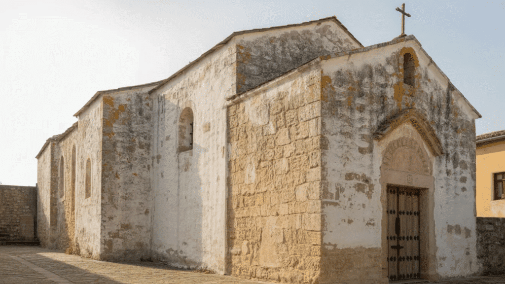 weathered ancient stone church with limewashed walls, arched windows, wooden door, and cross on roof under soft daylight