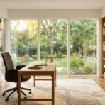 Modern home office with wooden desk, shelves with plants and pottery, large window overlooking garden