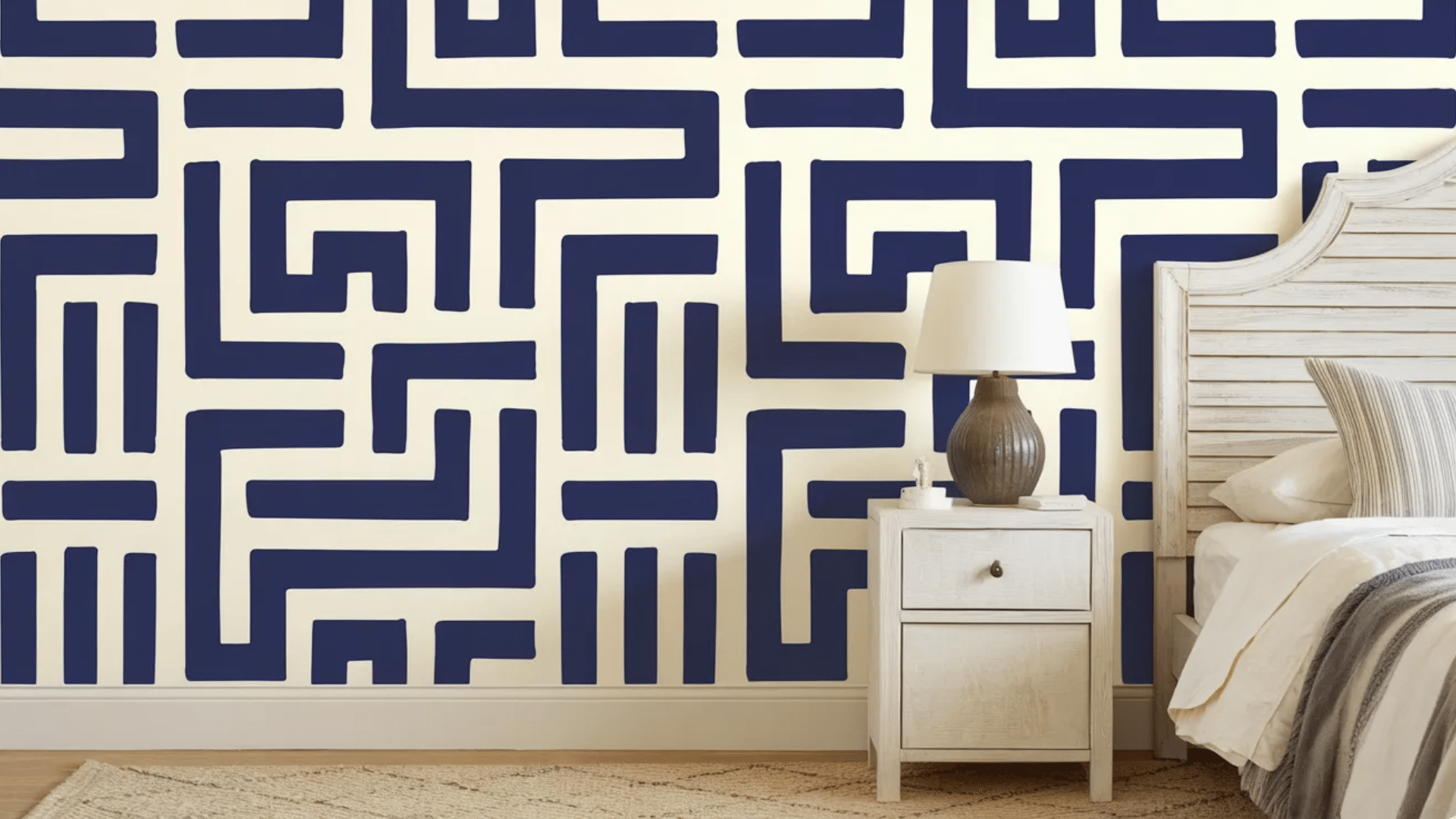 a bedroom with a bold, navy blue geometric maze-like wall design. The room features a simple white bed frame with a cozy white duvet and a patterned lamp placed on a nightstand. (1)