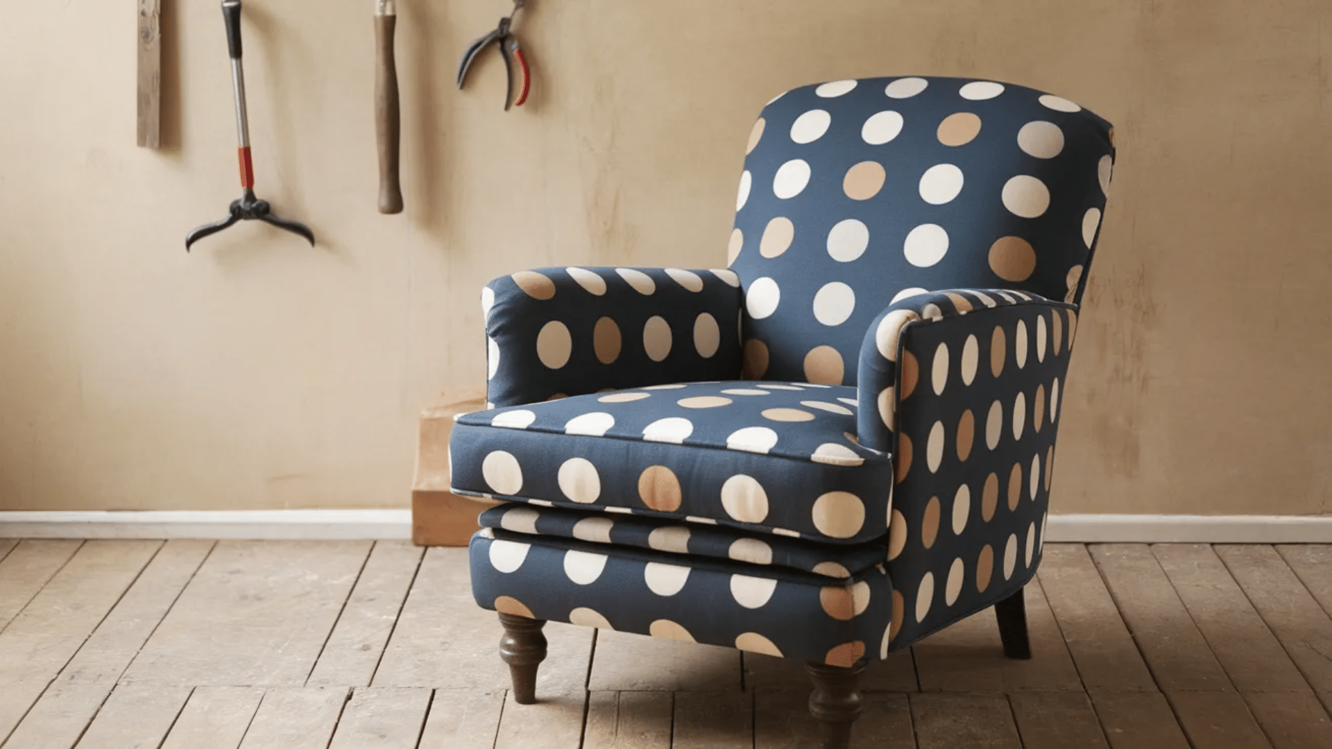 a blue armchair with a large polka dot pattern in white, beige, and light brown, placed on wooden flooring with gardening tools in the background.