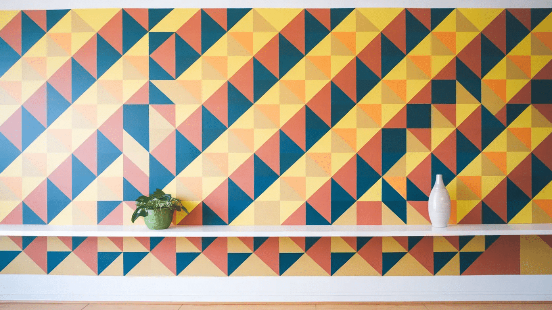 a colorful geometric wall featuring triangles in shades of yellow, orange, red, and teal, with a white shelf and a green potted plant on it.