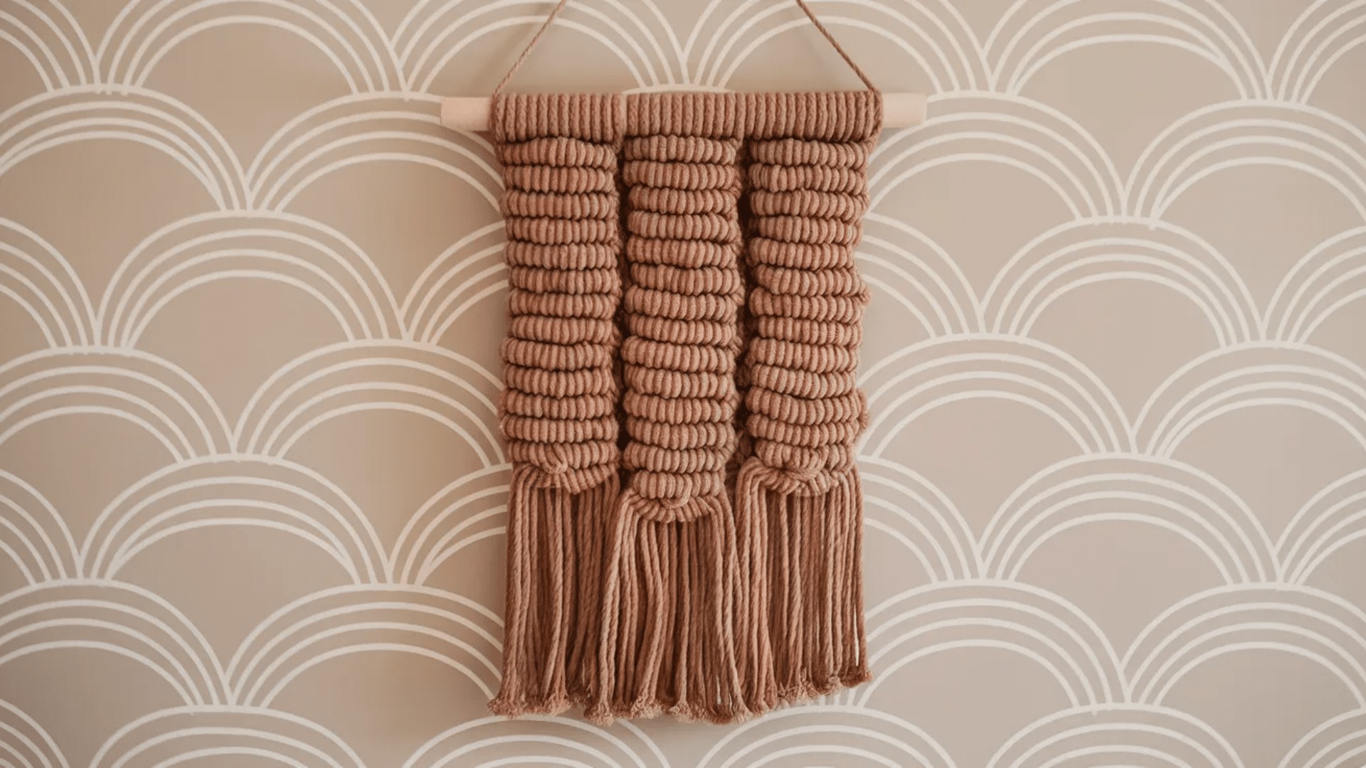 a macrame wall hanging in a tan color with knotted patterns and fringe tassels, displayed on a soft beige wall with white arched lines.