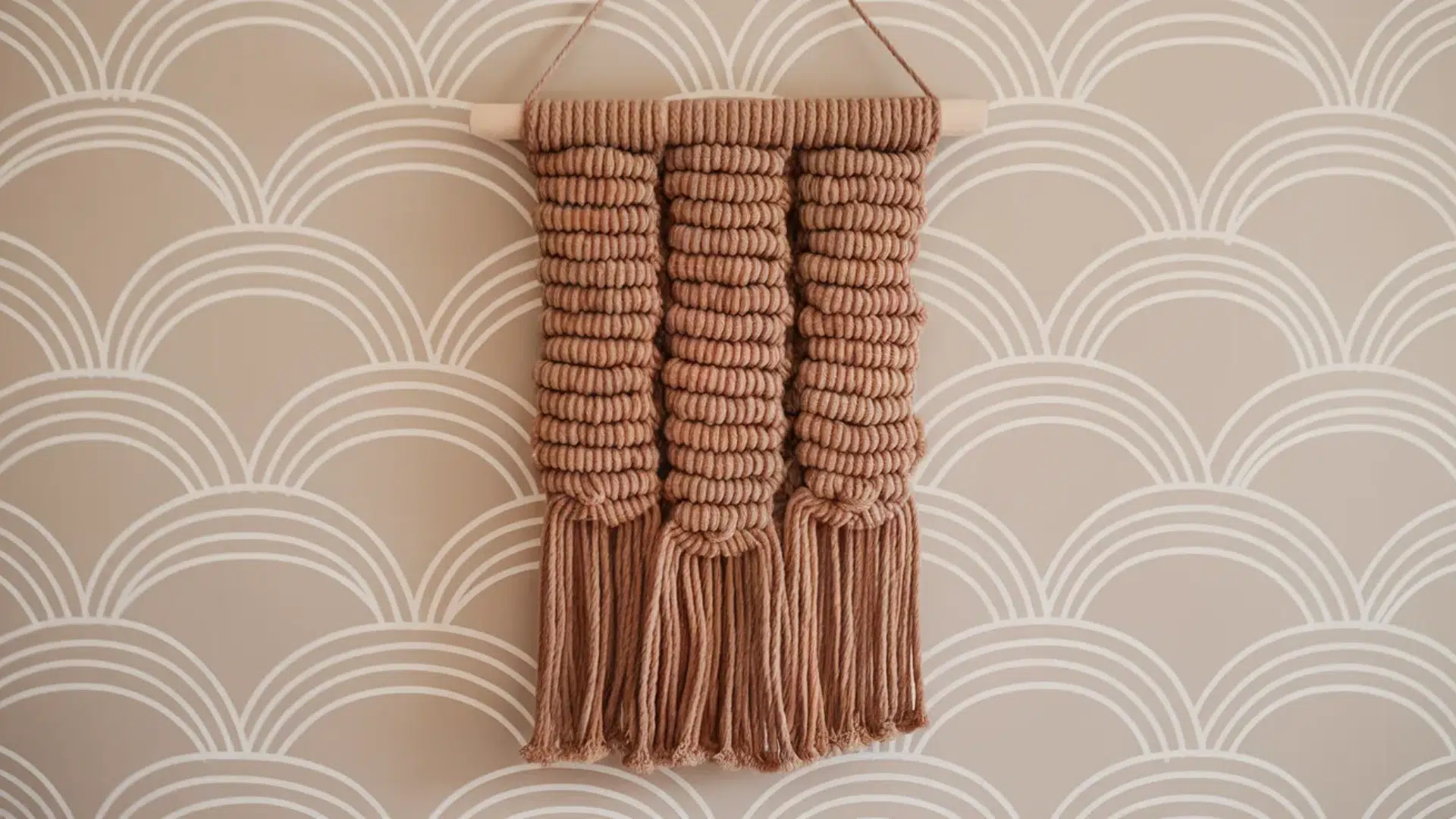 a macrame wall hanging in a tan color with knotted patterns and fringe tassels, displayed on a soft beige wall with white arched lines.