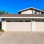 Beige two-car garage with brown shingled roof in residential neighborhood under clear blue sky