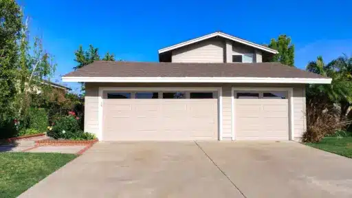 Beige two-car garage with brown shingled roof in residential neighborhood under clear blue sky